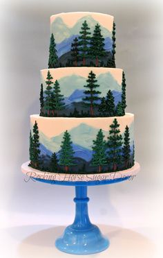 a three tiered cake decorated with trees and mountains is on a blue stand against a white background