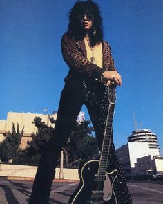 the man is posing with his guitar in front of some buildings and blue sky behind him