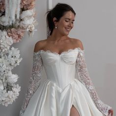 a woman in a white wedding dress with long sleeves and high slits standing next to flowers