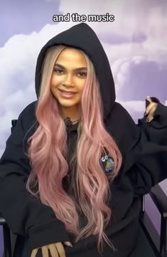 a woman with long pink hair sitting in front of a purple background and wearing a black hoodie