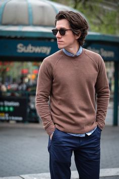 a man wearing sunglasses and a brown sweater is standing on the sidewalk with his hands in his pockets