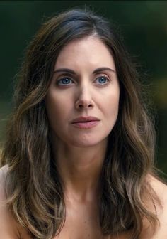 a woman with long hair and blue eyes looks off to the side in front of trees