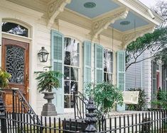 a white house with blue shutters and green plants on the front porch, next to a wrought iron fence