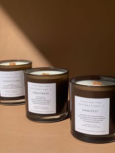 three candles sitting next to each other on top of a brown table with white labels