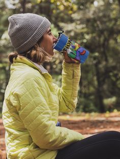 a woman sitting on the ground drinking out of a coffee cup with trees in the background