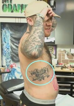 a man with a hat on his head has tattoos on his back and arm, while he sits in a chair