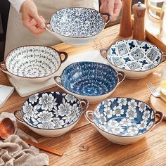 a person holding a bowl with six bowls in it on a wooden table next to plates and utensils