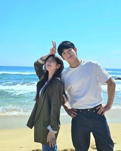 a man and woman standing next to each other on a beach with the ocean in the background