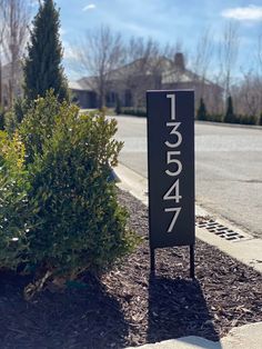 a black sign sitting on the side of a road next to a tree and bushes