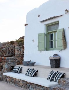 a white house with green shutters and striped pillows on the front porch, next to a stone wall