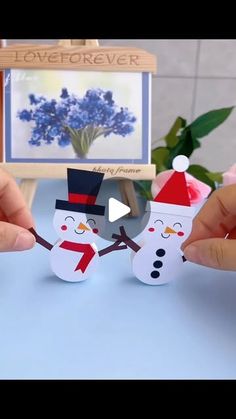 two hands holding up paper snowmen on top of a blue table with flowers in the background