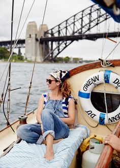 a woman in overalls sitting on the front of a sailboat with a bridge in the background