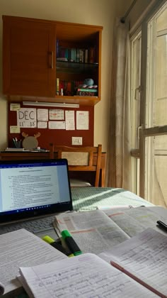 an open laptop computer sitting on top of a desk next to books and papers in front of a window