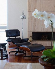 an eames chair and ottoman in front of a fireplace with flowers on the table