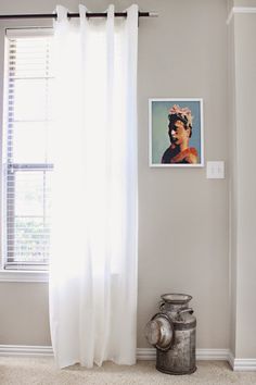 a window with white curtains and a painting hanging on the wall next to a trash can