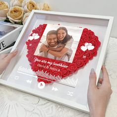 two hands holding a framed photo with roses in the shape of hearts and an arrow