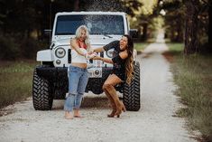 two women are shaking hands in front of a jeep on a dirt road with trees behind them