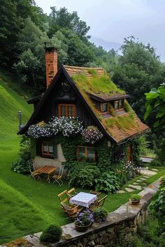 a small house with a green roof and flowers on the windows, surrounded by greenery