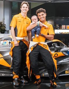 two men holding a baby in front of a race car