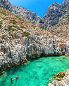 two people are swimming in the clear blue water near some mountains and cliffs on a sunny day