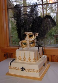 a three tiered cake with black feathers and masks on top, sitting in front of a window