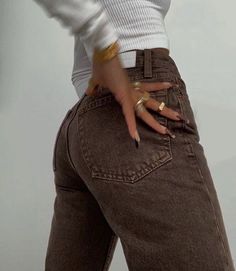 a woman with her hand on her hip wearing brown jeans and gold bracelets, standing in front of a white wall