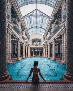 a woman sitting on the edge of a swimming pool in an indoor area with columns