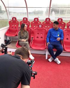 two people sitting on red seats in front of camera equipment, one person is filming the other