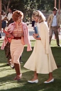 an old photo of two women talking to each other on the grass with people in the background