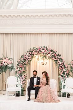 a man and woman sitting on chairs in front of a floral arch with pink flowers