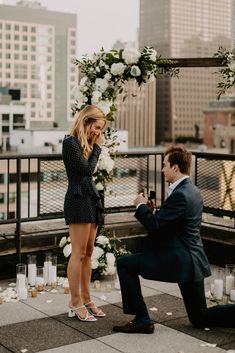 a man kneeling down next to a woman in front of a wedding arch with flowers on it