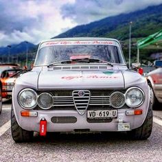 an old car is driving down the road with other cars behind it and mountains in the background