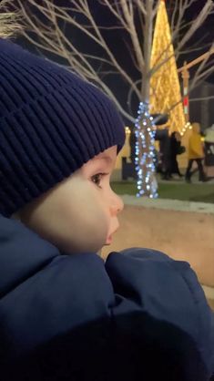 a little boy wearing a blue hat looking at the christmas tree in front of him