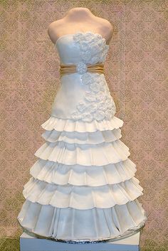 a dress made out of white ruffles and gold ribbon on display in front of a wall