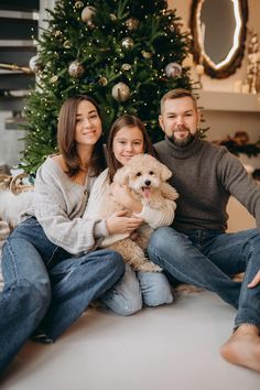 a man and woman sitting on the floor with a dog in front of a christmas tree