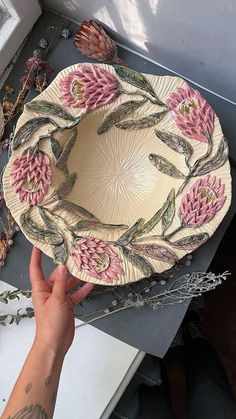 a hand holding a decorative plate with pink flowers and leaves on the bottom, in front of a window