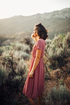 a woman in a pink dress standing on a hill looking up at the sky with her hands behind her head