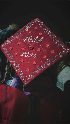 a red graduation cap with flowers and the words sold written in white on it, sitting among other people