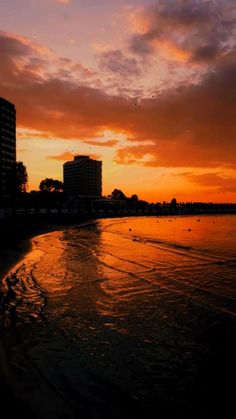 the sun is setting at the beach with buildings in the background and water on the shore