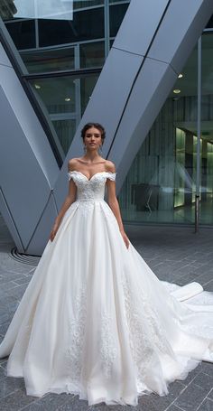 a woman standing in front of a building wearing a wedding dress with an off the shoulder neckline