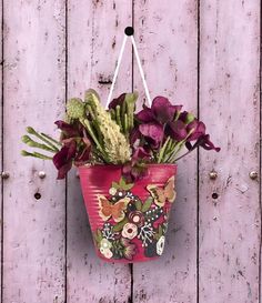 a pink flower pot hanging on a wooden wall with purple flowers and greenery in it