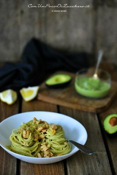 a white bowl filled with pesto pasta on top of a wooden table next to an avocado