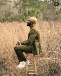 a man sitting on top of a wooden stool in front of tall grass and trees