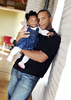 a man holding a baby in his arms while standing next to a brick wall and looking at the camera