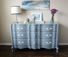 a blue dresser with flowers and pictures on the top, in front of a painting