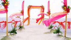 an outdoor ceremony set up with pink and orange sashes, flowers and grass on the beach