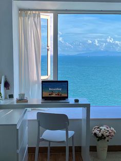 a laptop computer sitting on top of a white desk next to a window overlooking the ocean