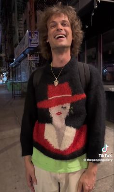 a man standing on the sidewalk wearing a sweater with a woman's face painted on it