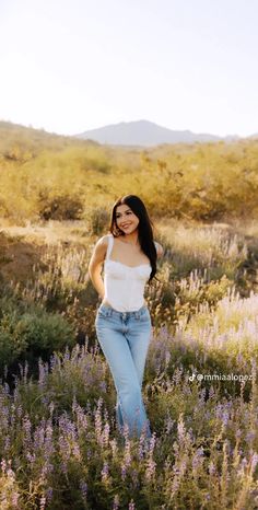 a woman standing in the middle of a field with purple flowers and mountains behind her