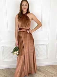 a woman standing in front of a white wall wearing a brown dress and holding a bouquet